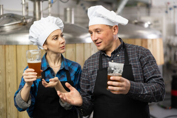 Woman and man skilled brewmasters standing on background of fermenters at brewery, checking quality of beer and discussing..