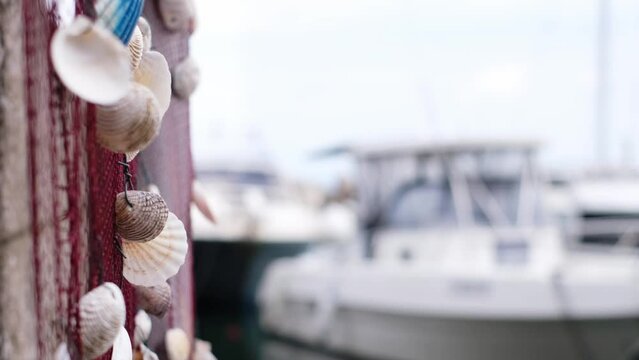 Beautiful selective focused sea shell decoration blowing by the wind. White sea shell pieces on a red fishing net. Slow motion natural sea vibes footage with defocused boats in the background. 4K