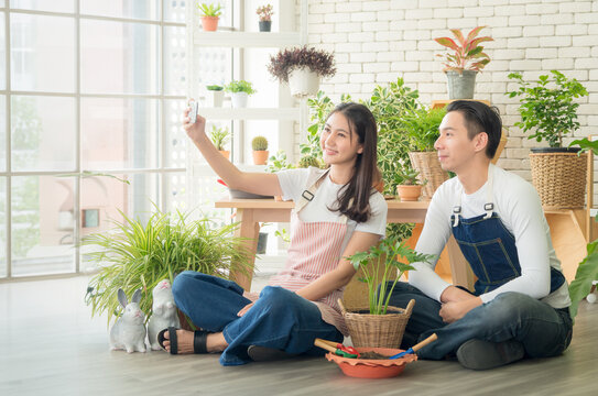 Couple gardener young Asian man woman two person sitting with floor smiling looking hand holding Smartphone selfie camera in calm work shop home plant white wall. hobby job happy and care concept