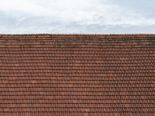 Old roof tiles in front of a cloudy sky. The red roofing is weathered and dirty. The overlapping...