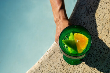 Close em um drinque refrescante com rodelas de limão e laranja sendo segurado por mão de mulher na beira da piscina em dia de sol.  - obrazy, fototapety, plakaty