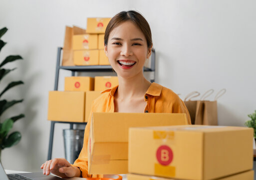 Startup small business, Young Asian woman holding packing boxes and checking online order on digital laptop for products to send to customers. working at the home office.