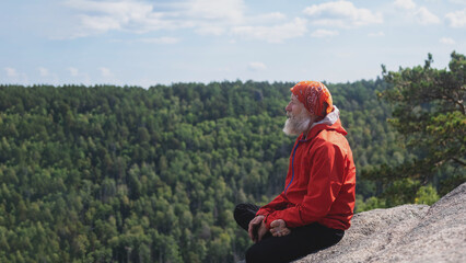 An elderly gray-haired man with a beard, a hipster traveler sits on a rock, relaxes, looking at nature from a height, after a hike to the top of the mountain.
