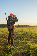  Man with gun stands on the autumn field in search of hunting object at dawn. Hunting period,...