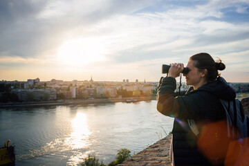 A young female traveler smiles, looks through binoculars and admires the view of the evening city and the river in the rays of the setting sun during her journey through Europe
