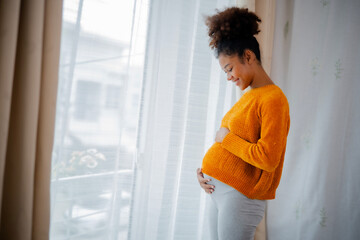 African American pregnancy woman in yellow sweater standing beside glasses door, embrassing and...