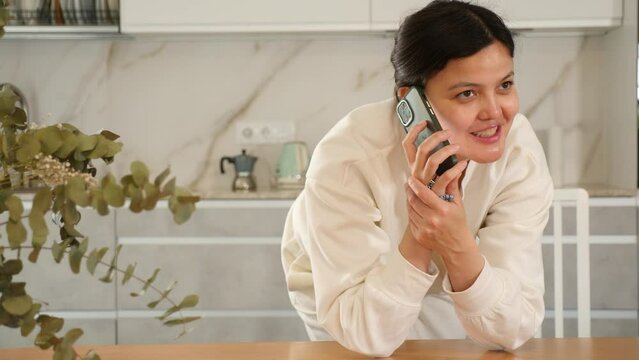Calm and cheerful young woman with dark hair dressed in comfortable white clothing, talking amiably on mobile phone while standing in cozy home kitchen 