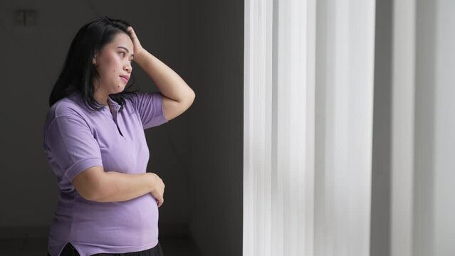 Overweight or pregnant Asian woman thinking of her problems standing near window 4K Footage of