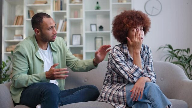 Married couple in casual attire having argument on grey couch at home in living room. Devastated african american woman crying while man desperately trying to find out reason of trouble