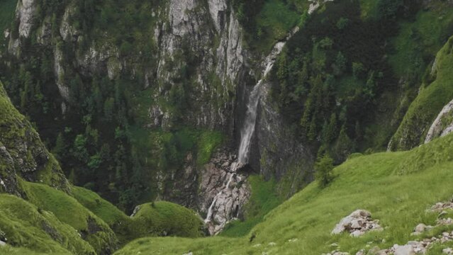 Slow motion static shot of a waterfall pouring into a mountain valley with an alpine landscape