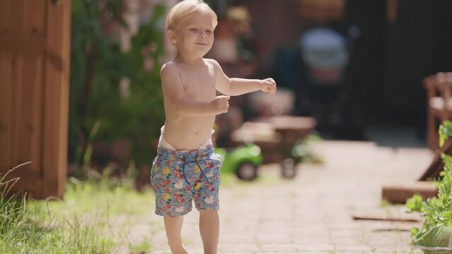 A little fair-haired bare-chested boy with his diaper sticking out of his shorts runs happily in the yard. Slow-motion. bokeh background.