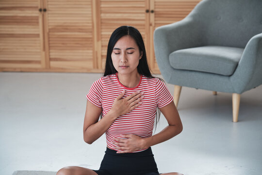 Asian Woman Practicing Yoga And Breathing Exercises In Morning, Sitting In Lotus Position With Eyes Closed Holding Hands On Chest And Stomach, Meditating At Home. Mindfullness And Mental Health