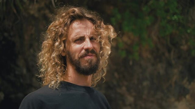Blinking in sunlight curly surfer with long hair on idyllic beach. Male hippie traveler on seaside. Caucasian man with beard on ocean shore. Enjoying summer vacation in tropical paradise. Travel.