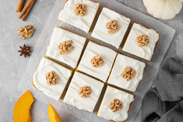 Spiced pumpkin cake with walnut and cream cheese frosting sliced into squares on a gray concrete background. Autumn dessert.