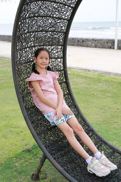 Portrait of Asian girl kid lying on circle rattan chair in the garden near beach.