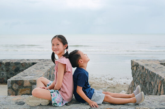 Adorable Asian young girl child and little boy kid sitting together at seacoast. Sister and brother sit back against each other on cement wall.