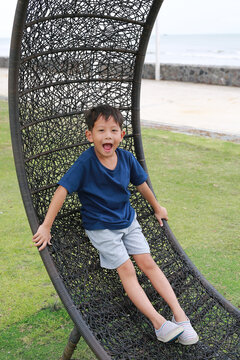 Portrait of Asian little boy lies on circle rattan chair in garden near coast.