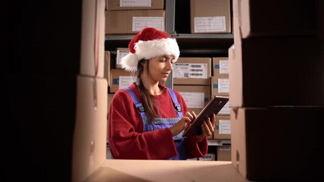 Warehouse worker in Santa hat uses digital tablet for checking stock, Christmas sale, on shelves standing cardboard boxes.