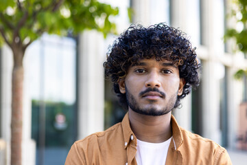 Close-up photo. Portrait, face of a young Indian man in an orange shirt standing outside on the street and looking seriously into the camera - obrazy, fototapety, plakaty