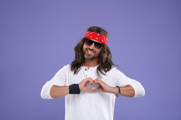 Stylish hippie man in sunglasses making heart with hands on violet background