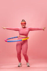 Full lenght portrait of young overweight woman dressed sporty training, warm up with hula hoop isolated on pink studio background.