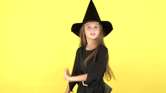 A little girl in a Halloween witch costume dances and shows emotions on a yellow studio background