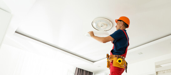 A young man stands on a stepladder and installs a large chandelier on the ceiling