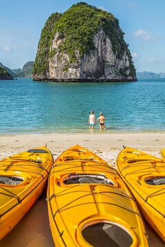 Kayaks lined up on a beach in front of two people standing the water and a large mountain at Ha Long Bay in Vietnam