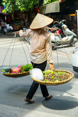 A street seller wearing an Asia reed hat balancing produce over their shoulder walking down the street in Hanoi in Vietnam