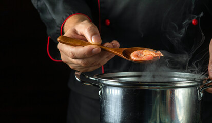 The chef prepares delicious shrimp in a pan in the kitchen. The concept of a delicious scampi lunch for a restaurant. Spoon in the cook hand wis prawn - obrazy, fototapety, plakaty