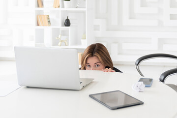 Fototapeta premium Hysterical business woman hiding behind desktop and looking at laptop in the office. Business crisis and bankruptcy concept