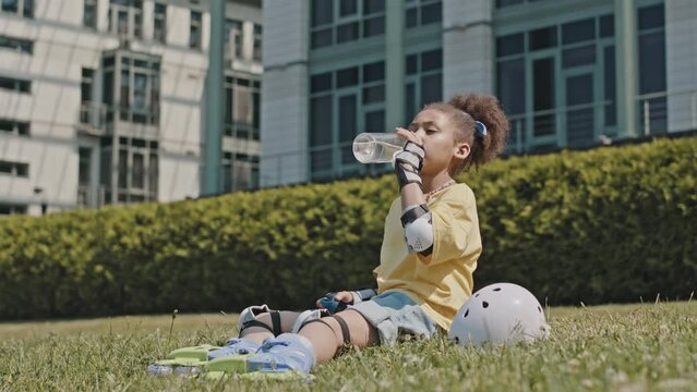 Tilt up shot of little African American girl in roller skates resting outdoors on green grass and drinking water from bottle