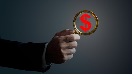 Fototapeta premium Businessman is using magnifying glass and searching for money against dark background.