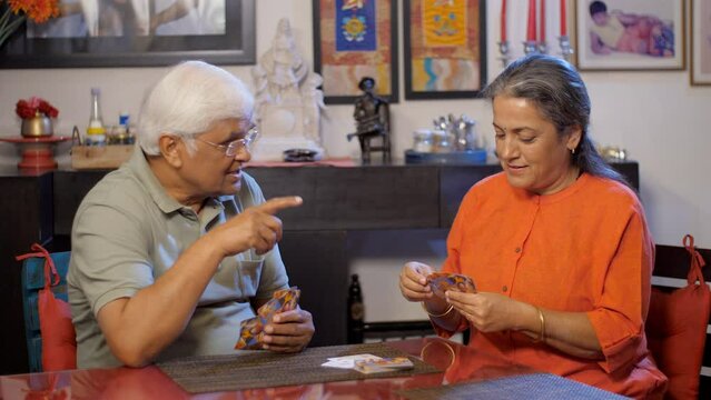 An aged Indian couple is playing a game of cards at home - leisure time  passing free time  relaxed lifestyle. Two happy senior people having fun playing cards while sitting at a dining table - a r...