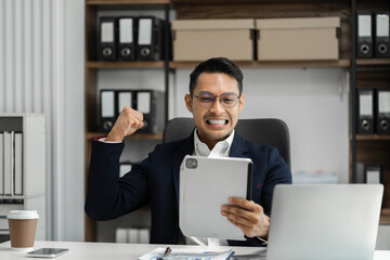 Fototapeta na wymiar Happy young businessman looking at successful excited tablet sitting at desk raising hands in yes gesture celebrating business success.