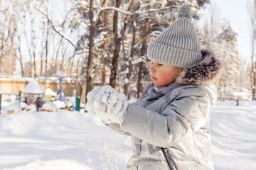 Happy child on winter walk. Little girl in warm suit plays in park and enjoys sunny day in winter, she stands on snowdrift and plays snowballs.