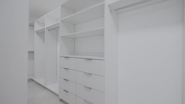 Large spacious empty walk-in closet with white cabinets, drawers, and holders for hangers. Beautiful wardrobe for placing clothes and shoes. Interior design concept