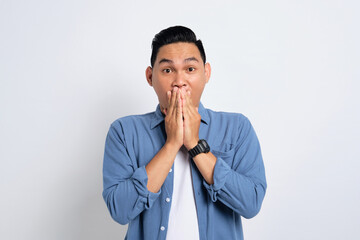 Surprised young Asian man in casual shirt covering mouth with hand, looking at camera with amazed facial expression isolated on white background