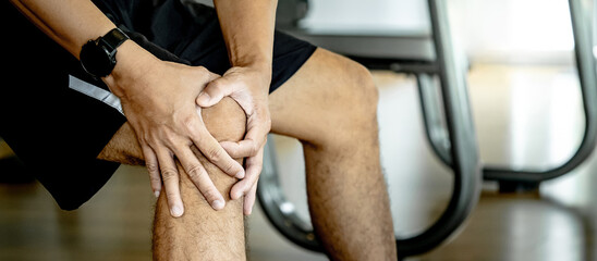 Sportive man feeling knee pain or rheumatoid arthritis while sitting on workout bench in fitness...