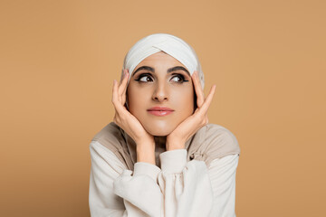 portrait of positive and dreamy muslim woman in hijab looking away with hands near face on beige