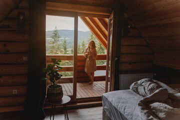 Young relaxed cheerful woman enjoying the morning and nature on the cabin balcony at sunrise with a cup of tea - obrazy, fototapety, plakaty