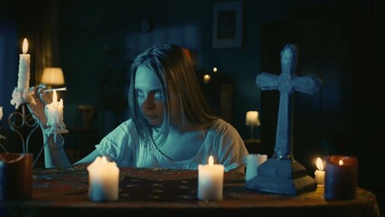 Shot capturing a young woman sitting at the table, surrounded by candles, crosses and beads,...