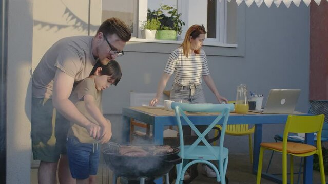 Father is teaching his son how too work with the grill while mother sets the table.