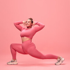 Full body portrait of young plus size sporty woman in pink top and legging training, stretching legs holdings hands near head isolated studio background.