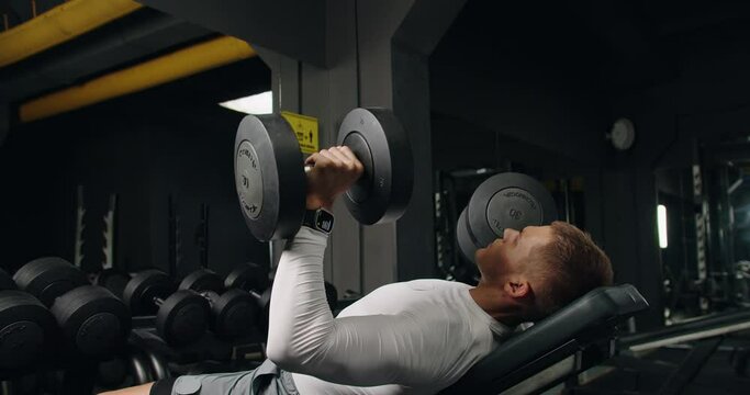 Athletic man has chest workout and doing bench press with heavy dumbbells in gym. Muscular male in sportswear exercising with lifting weight at fitness club. 