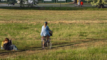 Park life: people relax and have fun in a green park with trees. A woman in a blue shirt rides a bicycle, two female friends are sitting on the grass, a warm summer evening