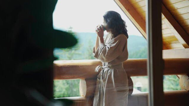 A young woman in a bathrobe stands on the terrace of a country log house and enjoys her morning coffee and fresh mountain air.