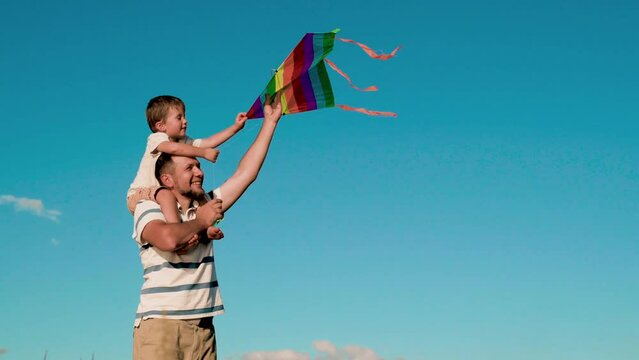 A small son sits on his father's neck and launches a kite. Papa rolls his son around his neck in the summer against the sky playing with a kite in the wind