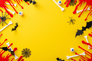 Fototapeta na wymiar Unconventional Halloween theme. Top view perspective of themed adornments, spooky blood smears, bones, eyeballs, insects, spiders, spiderweb, bats on yellow base, vacant frame for text or ads