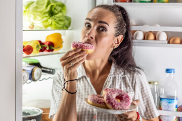 Hungry brunette in pajamas enjoys sweet donuts late at night by the open refrigerator